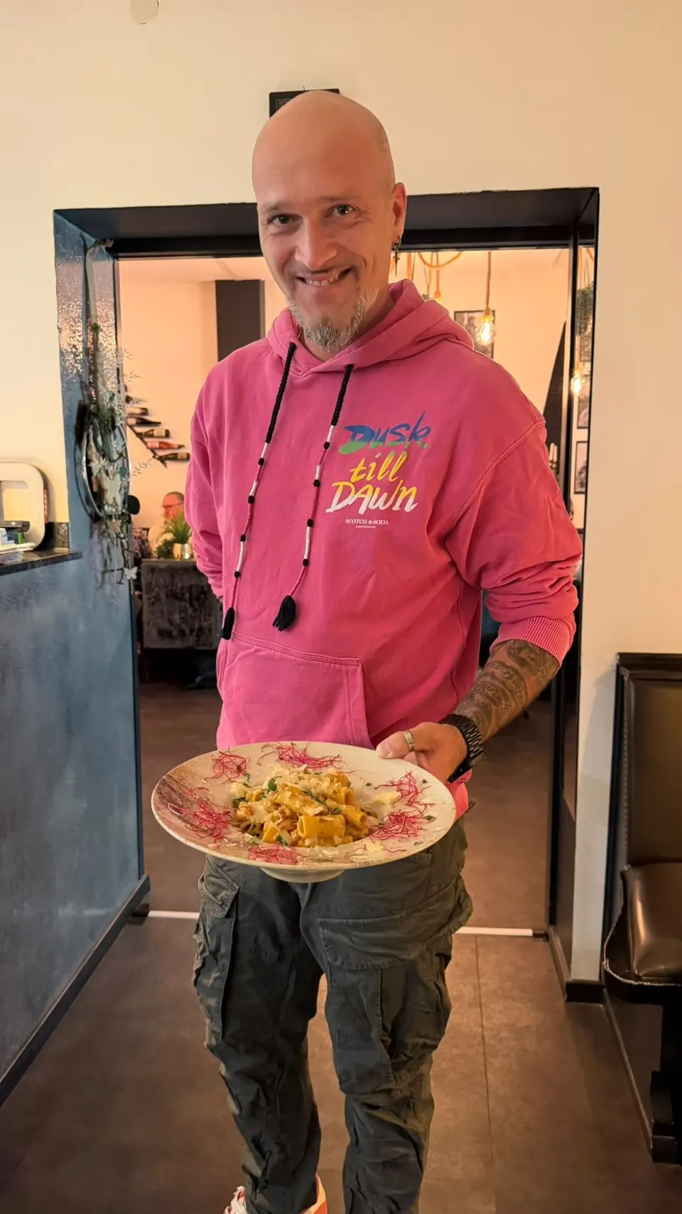 Gastgeber Niko lächelnd mit einem Teller Rigatoni in Tomatensauce mit Parmesan